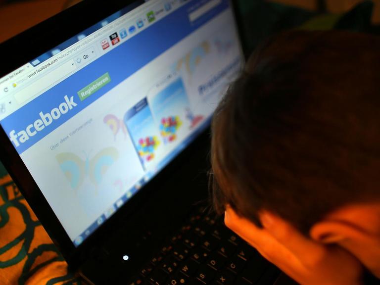 Ein Mensch sitzt mit in den Händen vergrabenem Gesicht vor einem Bildschirm, auf der Facebook angezeigt wird.