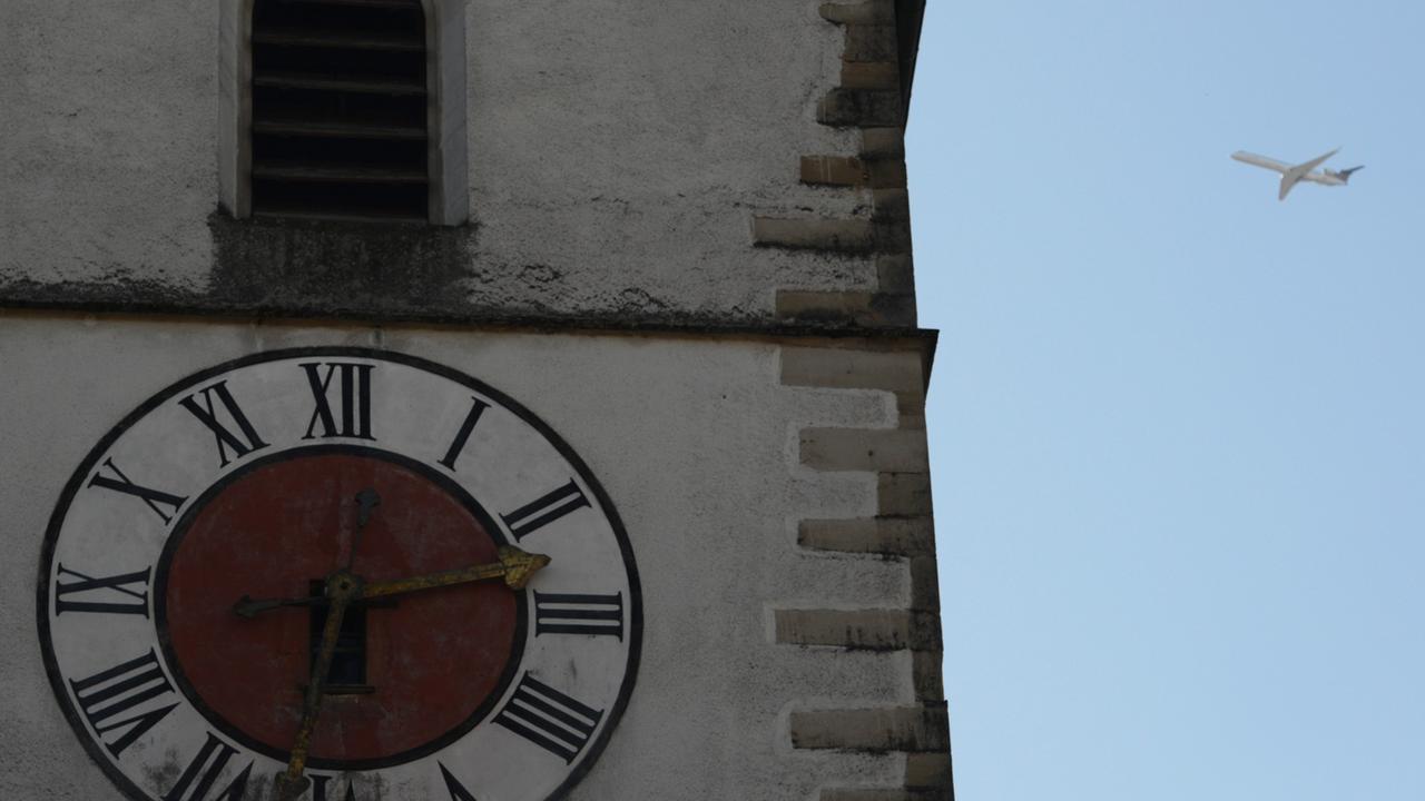 Die Uhr eines Kirchturms in Großaufnahme, daneben weit hinten ein Flugzeug im Landeanflug vor blauem Himmel.