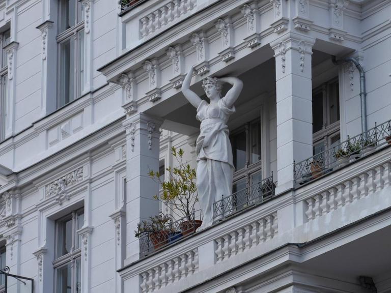 Blick auf eine verzierte Altbaufassade mit Balkon, auf dem eine Frauenfigur als Stützpfeiler zu sehen ist