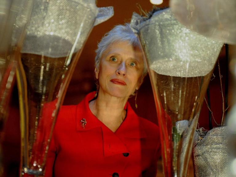 Schriftstellerin Sara Paretzky in rotem Kleid, im Vordergrund sehr groß zwei Sektgläser, in deren Mitte ist die Autorin zu sehen.