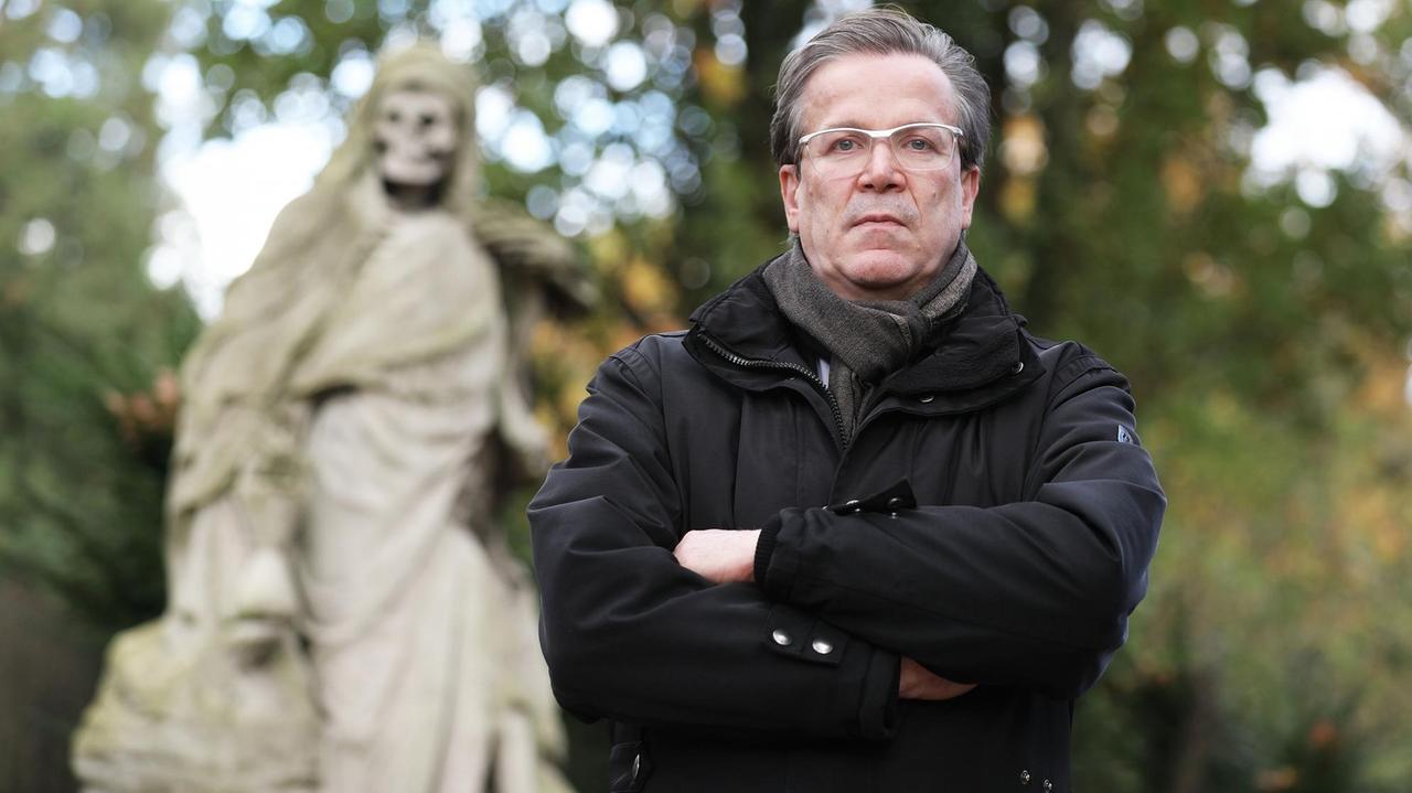 Christoph Kuckelkorn, Bestattungsunternehmer und Karnevalspräsident, steht auf einem Friedhof, links hinter ihm ein Grab mit einer Statue, die den Tod darstellt