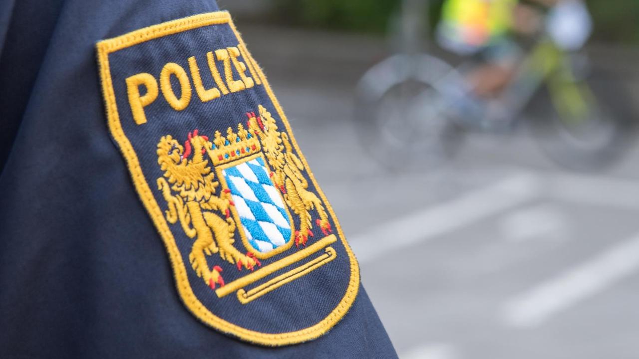 Das Logo der bayerischen Polizei ist auf dem Arm einer Uniform zu sehen, im Hintergrund verschwommen ein Fahrrad.