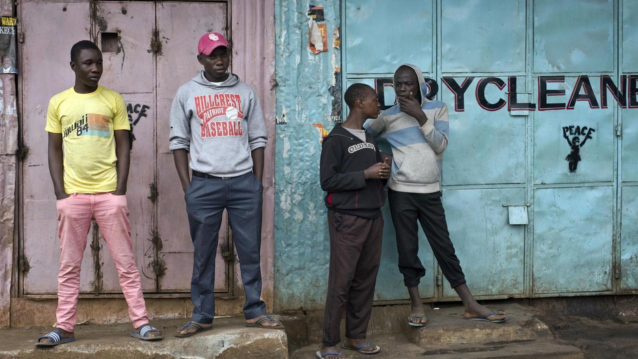 Die vier jungen Männer stehen nebeneinander vor zwei geschlossenen Läden und unterhalten sich. Auf einem der Läden steht "Drycleaner".