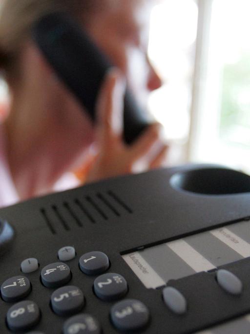Ein Telefon im Vordergrund. Im Hintergrund eine Frau, die einen Hörer ans Ohr hält.