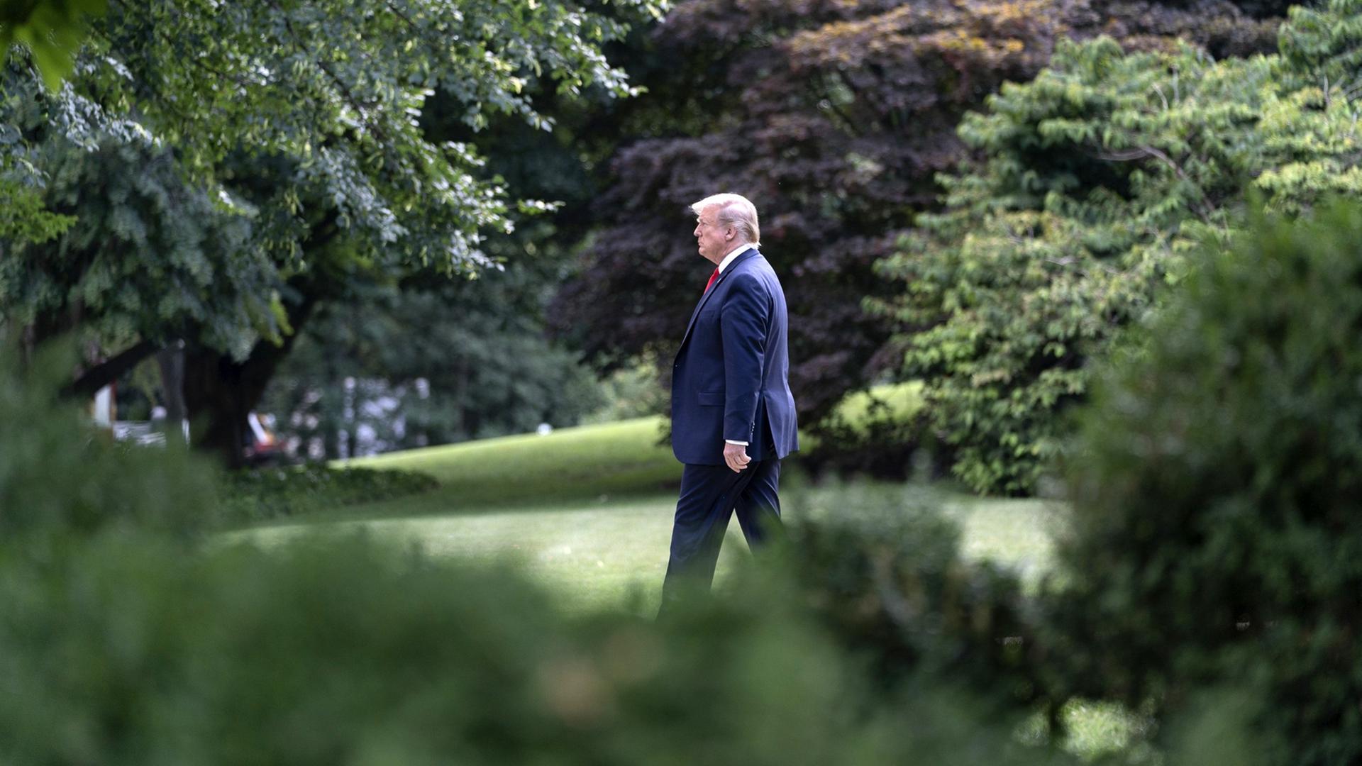 US Präsident Donald Trump geht alleine eingerahmt von Bäumen über einen Rasen.