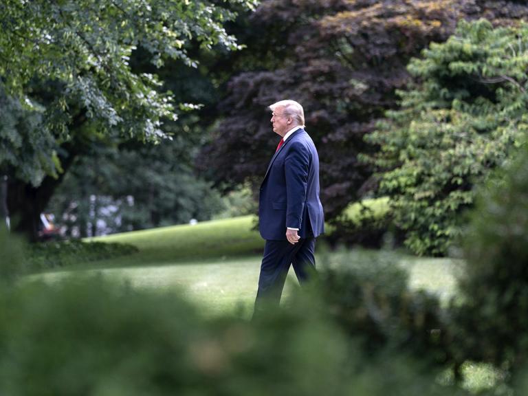 US Präsident Donald Trump geht alleine eingerahmt von Bäumen über einen Rasen.