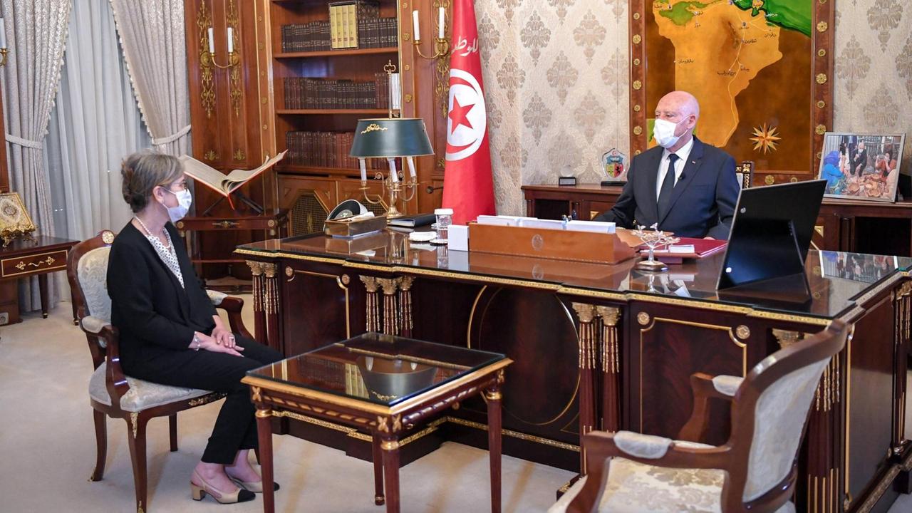 Der tunesische Präsident Kais Saied sitzt in Anzug und mit Mund-Nasen-Schutz an seinem Schreibtisch, dahinter eine Landkarte Tunesiens und die Landesflagge. Auf dem Besucherstuhl sitzt eine westlich gekleidete ältere Frau, ebenfalls mit Mund-Nasenschutz.