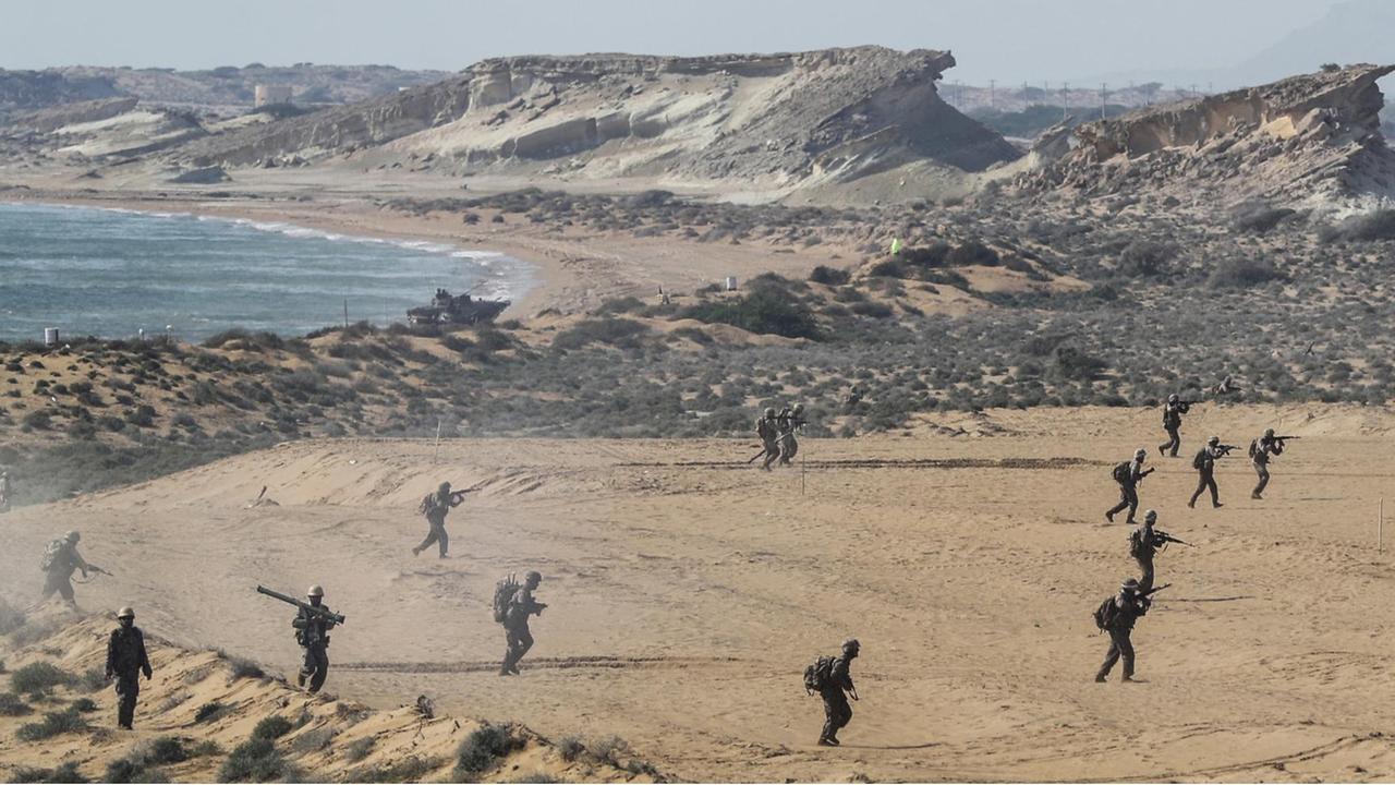 Blick aus der Vogelperspektive auf mehrere Soldaten an einem Strand am Meer. Man sieht sie als kleine schwarze Striche.