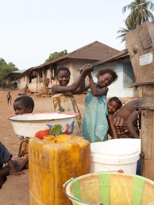Szene aus der Stadt Makeni, im Nordosten von Sierra Leone: Kinder pumpen sauberes Wasser aus einem Brunnen in Schüsseln und Kanister. Links daneben steht eine Frau in einem bunten Rock.