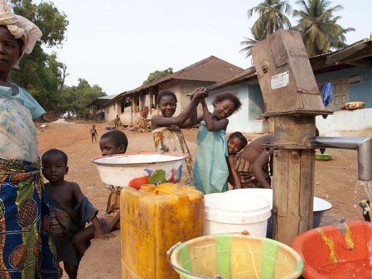 Szene aus der Stadt Makeni, im Nordosten von Sierra Leone: Kinder pumpen sauberes Wasser aus einem Brunnen in Schüsseln und Kanister. Links daneben steht eine Frau in einem bunten Rock.