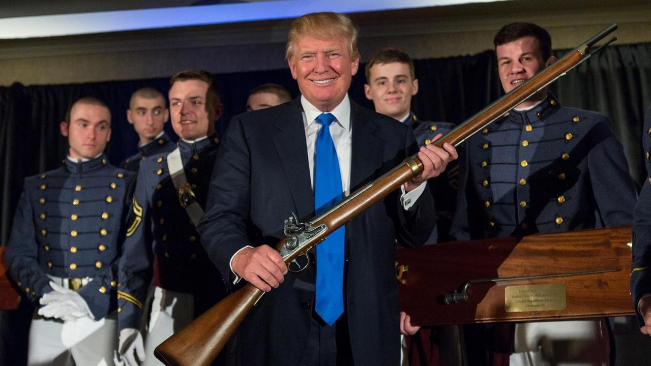 Donald Trump hält in einem Militär College stolz eine Waffe in die Höhe.