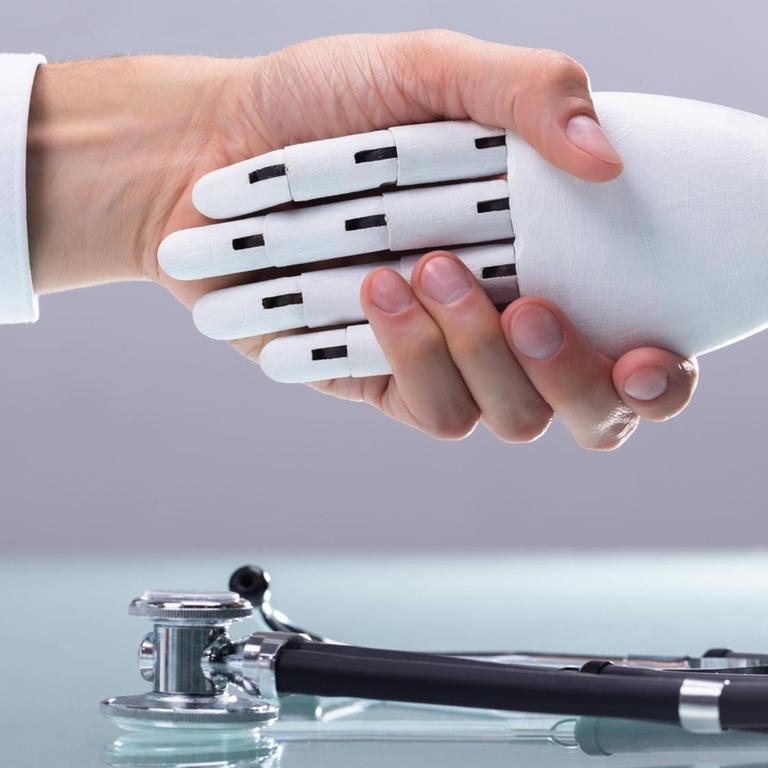 Handschlag einer menschlichen und einer Roboterhand, auf dem Tisch darunter liegt ein Stethoskop