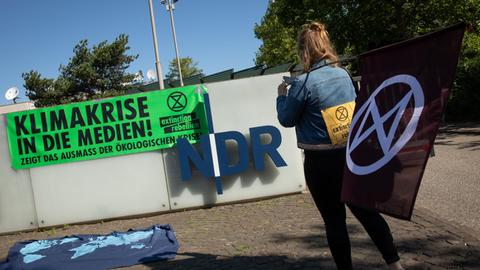 5.06.2020, Hamburg: Eine Aktivistin der Umweltschutzbewegung Extinction Rebellion (XR) steht während eines Protests mit einer Flagge vor dem Eingang zum Norddeutschen Rundfunk (NDR), an dessen Mauer ein Banner mit der Aufschrift «Klimakrise in die Medien! Zeigt das Ausmaß der ökologischen Krise» hängt. Mit einer Blockade vor der Einfahrt zum NDR-Gelände in Hamburg-Lokstedt haben die Umweltaktivisten den Sender aufgefordert, in seiner Berichterstattung den Begriff Klimawandel durch Klimakrise zu ersetzen. Foto: Christian Charisius/dpa
