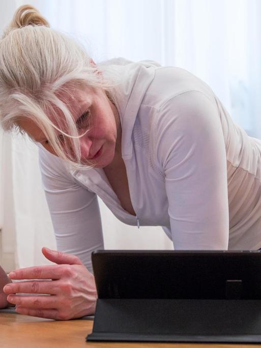 Eine ältereFrau macht in einer Wohnung Übungen und schaut dabei auf ein Tablet.
