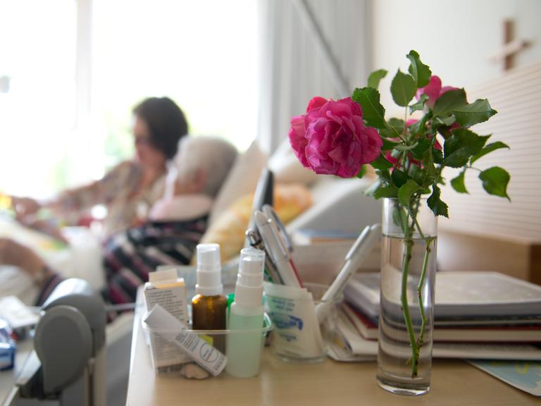 Im Vordergrund eine Rose, im Hintergrund ein Krankenbett mit einer alten Frau und einer jüngeren am Bett.