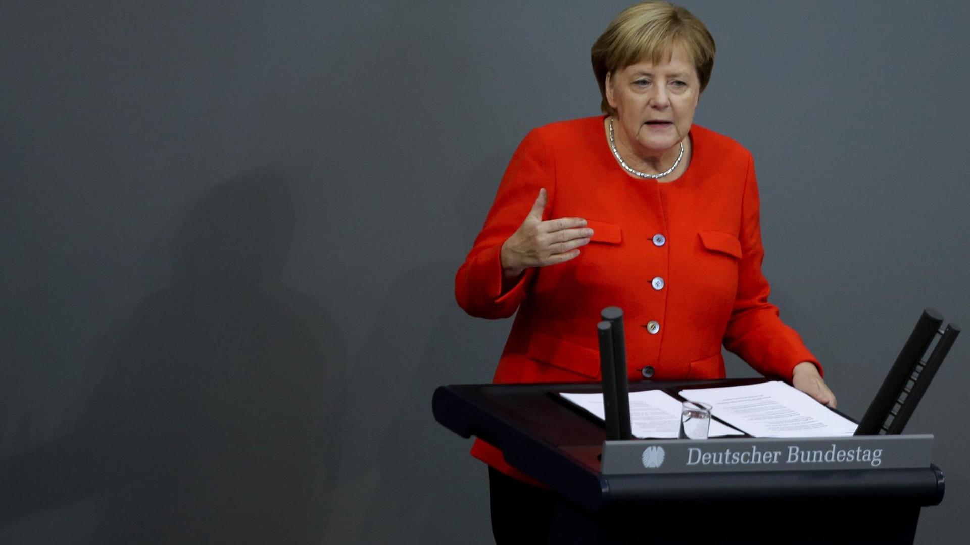 Bundeskanzlerin Merkel steht in einem roten Blazer hinter einem Rednerpult mit dem Schriftzug Deutscher Bundestag und gestikuliert mit der rechten Hand.