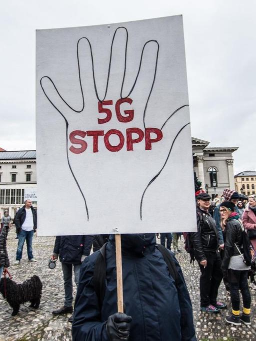 Ein Mensch verdeckt sein Gesicht mit einem großen Demoschild, auf dem eine Hand aufgemalt ist und '5G STOPP' steht.