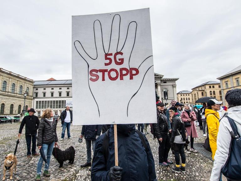 Ein Mensch verdeckt sein Gesicht mit einem großen Demoschild, auf dem eine Hand aufgemalt ist und '5G STOPP' steht.