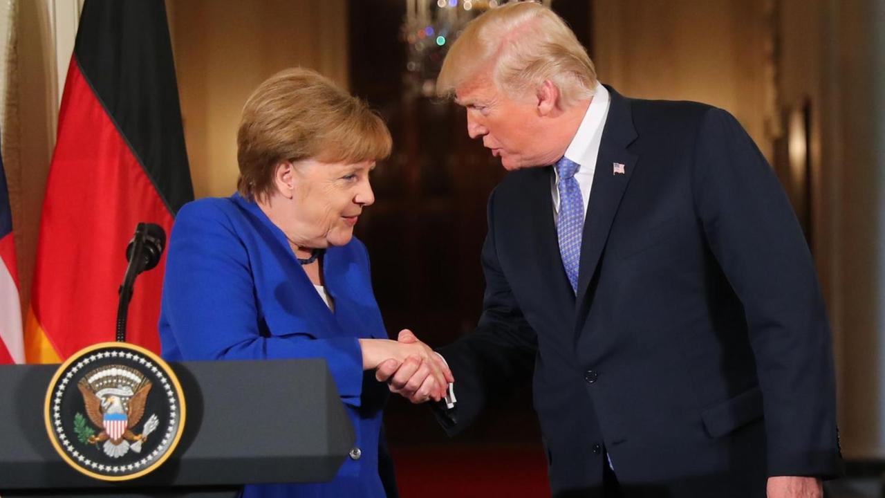 Beide beugen sich zueineander hin und schütteln die Hände. Trump sagte etwas, Merkel hört lächelnd zu. IM Vordergund ein Sprechpult mit Mikrofon und dem Präsidenten-Wappen.