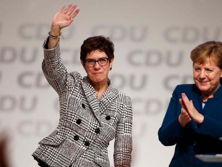 Annegret Kramp-Karrenbauer winkt auf dem Parteitag den Delegierten zu, neben ihr applaudiert Angela Merkel