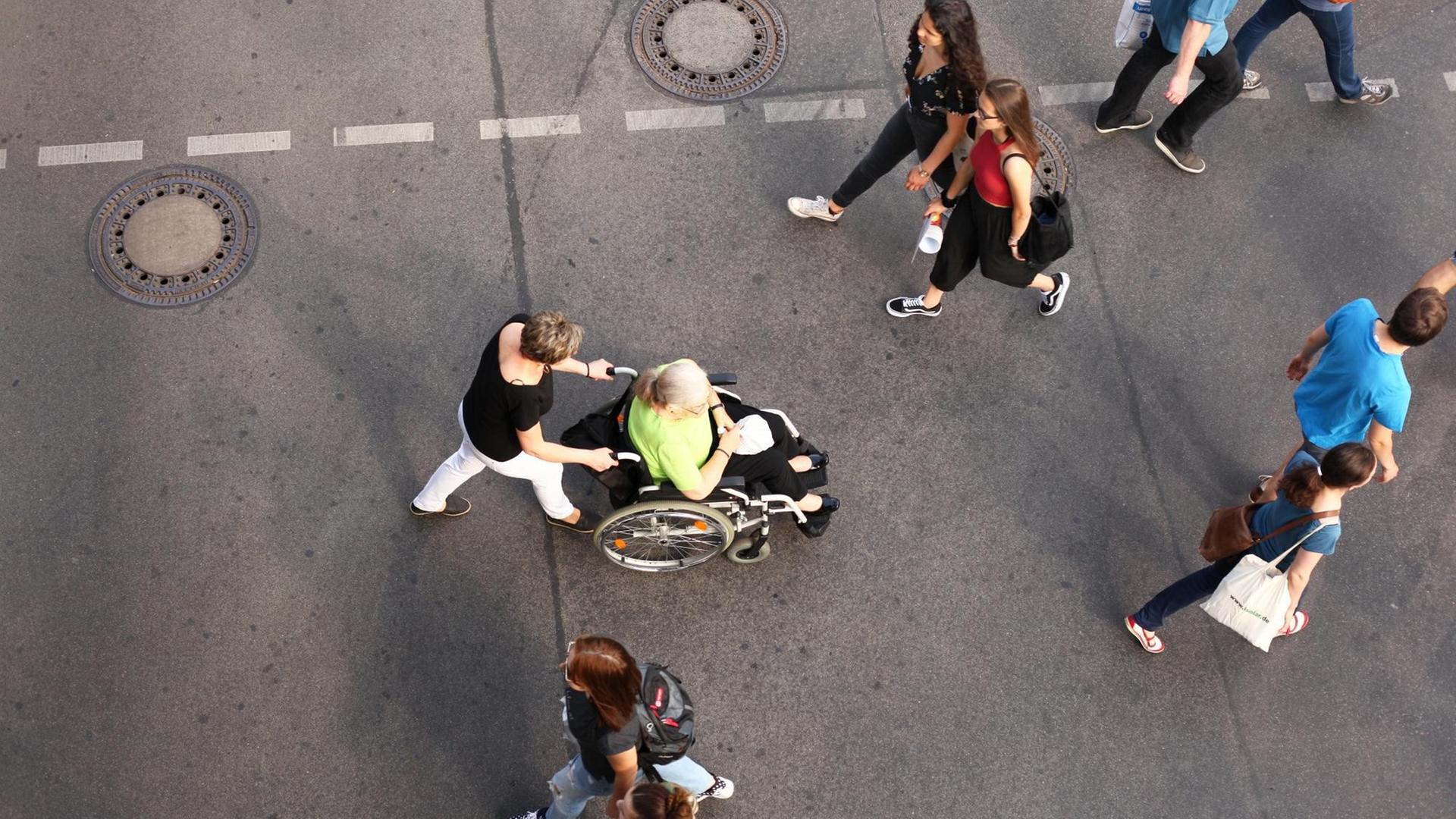Das Bild zeigt eine Straße von oben mit Menschen, in der Mitte eine Frau im Rollstuhl.