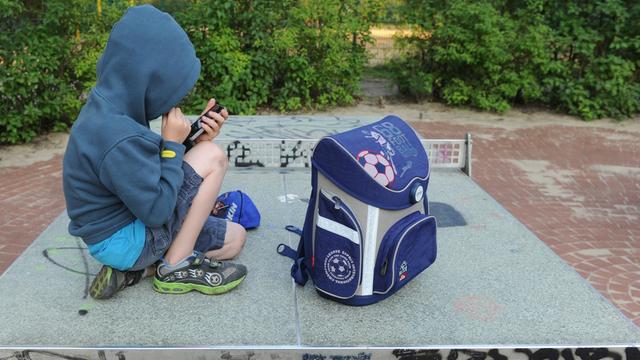 Ein Junge sitzt mit seinem Schul-Ranzen auf einer Tischtennis-Platte und spielt auf einem Handy.