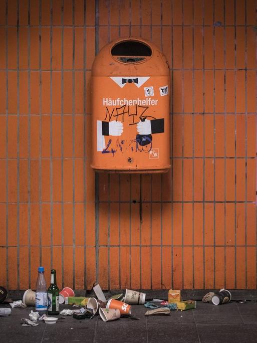 Ein orangener Mülleimer an einer orang gekachelten Wand in Berlin. Darauf steht Häufchenhelfer. Darunter liegt verschiedenartiger Abfall.