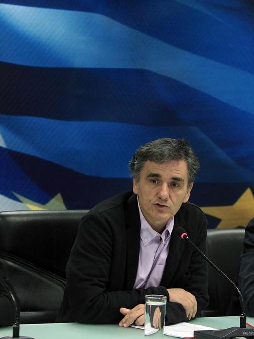 Dombrovskis, Tsakalotos und Stathakis sitzen an einem Pult. Tsakalatos spricht in ein Mikrofon. Die drei sitzen vor einer Wand, auf der die Fahnen Griechenlands und der EU zu sehen sind.