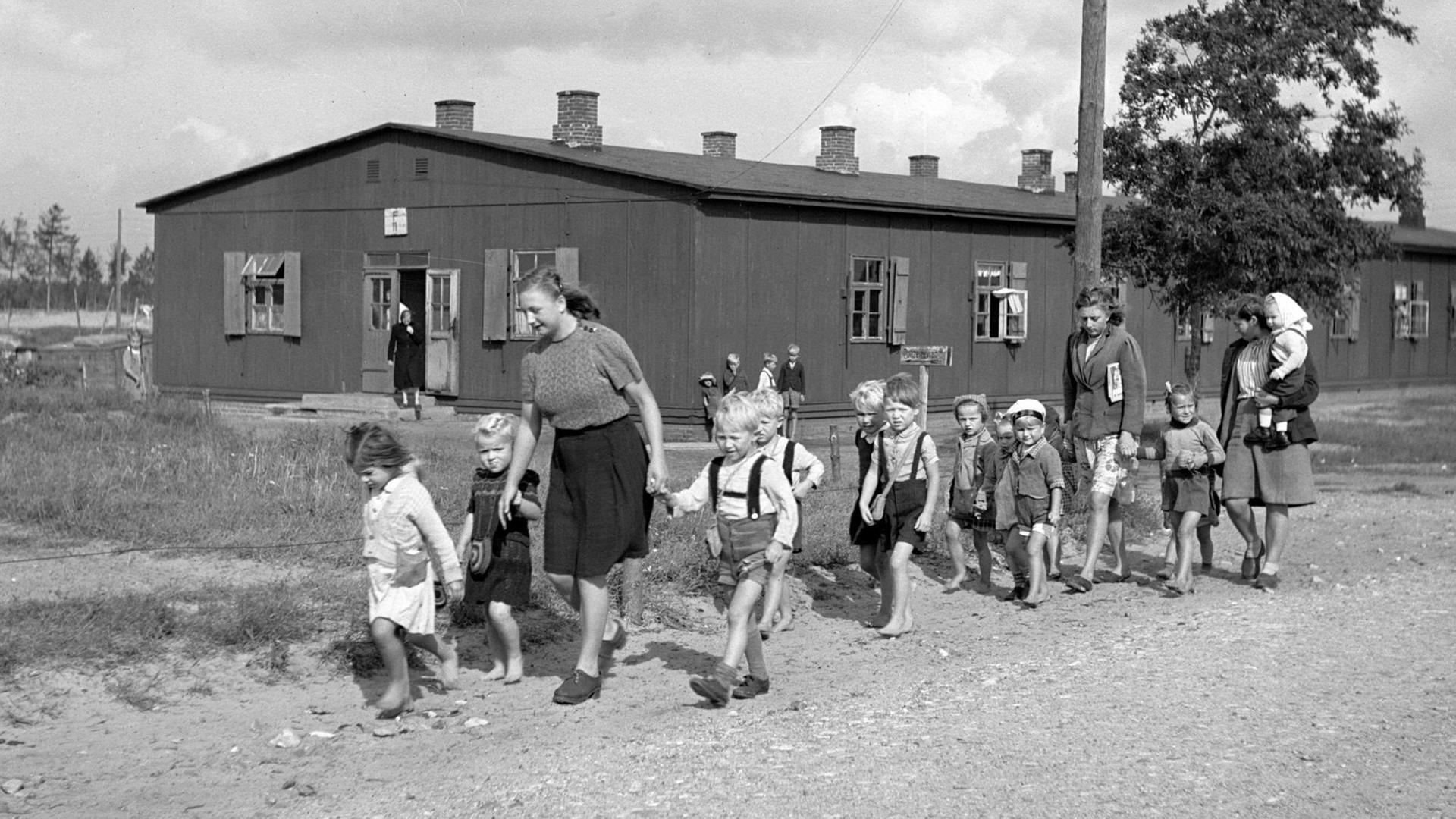 Eine Gruppe von Kindern, begleitet von zwei Frauen, läuft einen Weg entlang. Im Hintergrund sind Holzhäuser zu sehen.