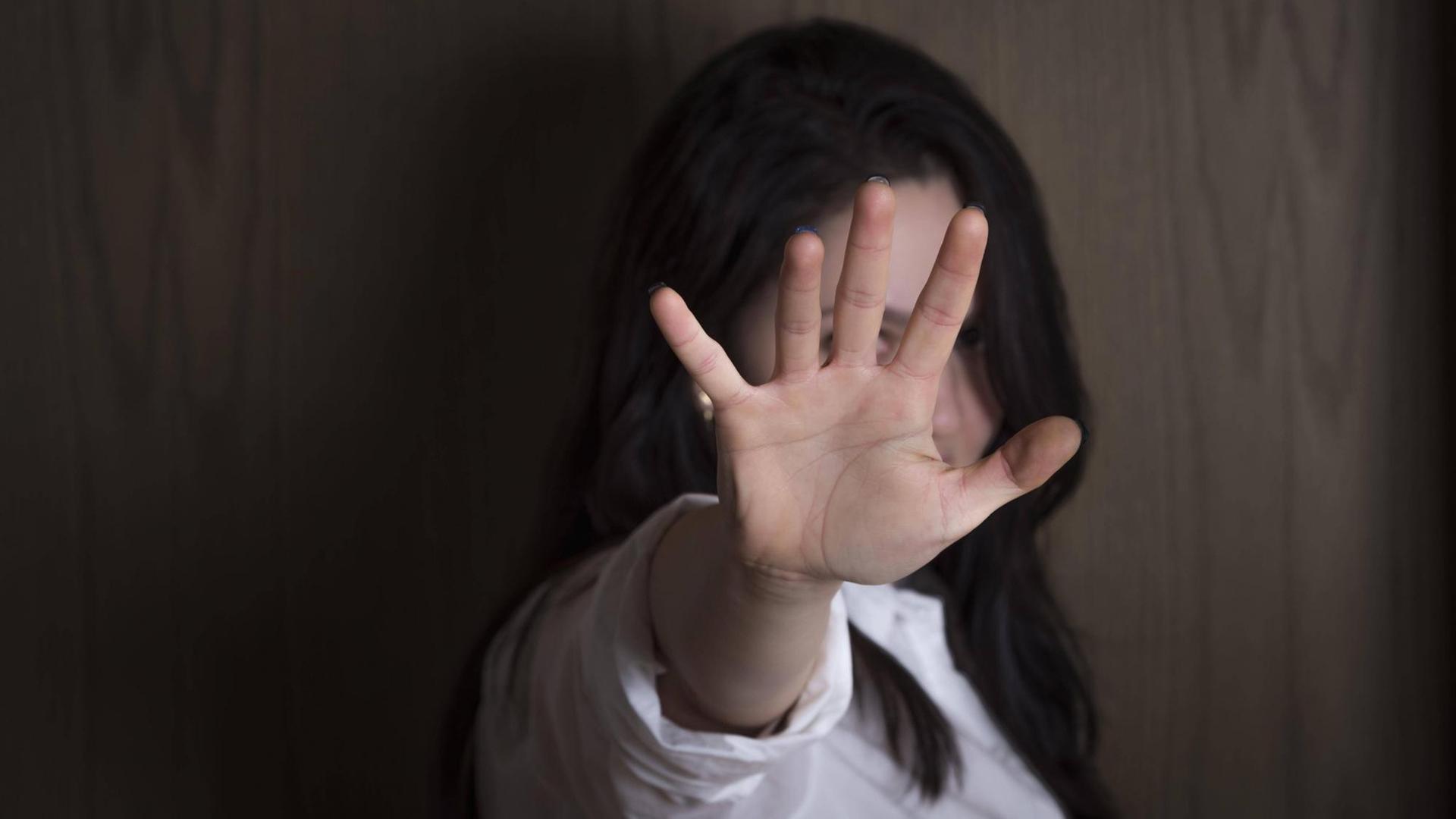 Eine Frau vor einer Holzwand hat ihre Hand zu einer Stopp-Geste ausgestreckt, und verdeckt so ihr Gesicht.