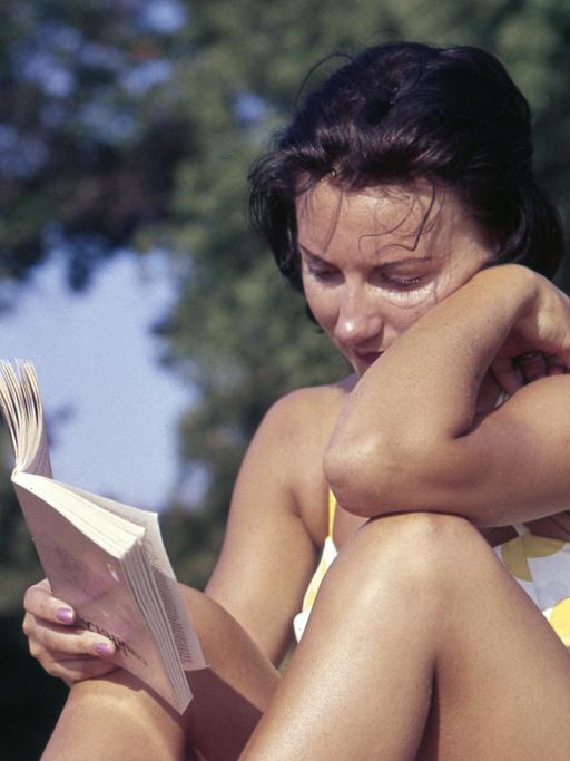 Eine Frau sitzt in einem Bikini auf einer Wiese und liest ein Buch.