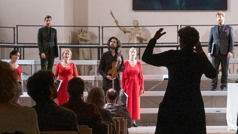 Ein Geiger steht neben Sängerinnen in roten Kleidern, im Vordergrund steht die Dirigentin, die nur als Schattenriss erkennbar ist.