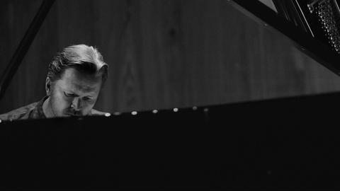 S/W Bild. Das Gesicht eines Mannes ist hinter einem Flügel am äußeren linken Rand zu sehen. Der Gesichtsausdruck konzentriert, der Blick gesenkt.