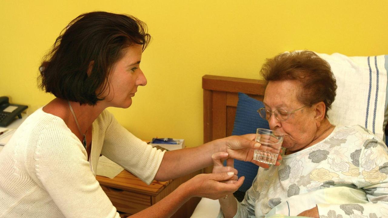 Eine Pflegerin reicht einer bettlägerigen Seniorin ein Glas Wasser.
