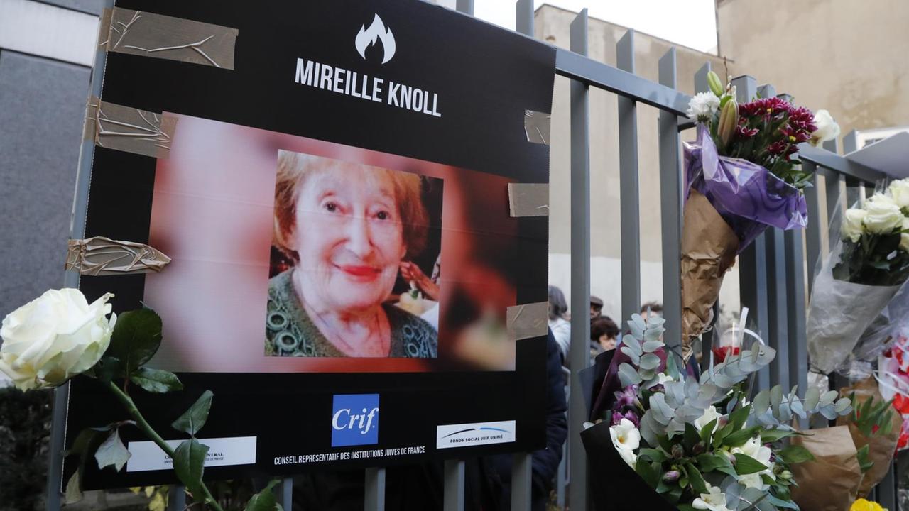Das Bild zeigt ein Foto von Mireille Knoll und Blumen am Zaun vor dem Haus, wo sie in Paris gelebt hat und ermordet wurde.