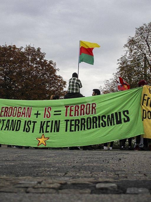 Demonstranten halten Banner hoch. Auf dem Banner, das im Zentrum des Bildes zu sehen ist steht: "Erdogan + IS = Terrorismus. Widerstand ist kein Terrorismus". Einer der Demonstranten hält eine kurdische Flagge hoch.