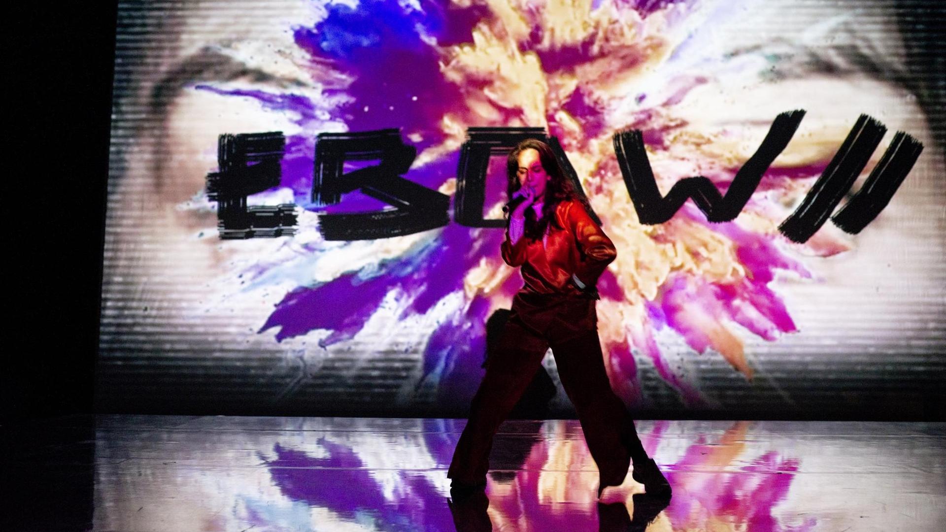 Die Künstlerin Ebow performt vor einer riesigen Leinwand. Darauf ist eine Farbexplosion zu sehen und ihr Name in schwarzen Buchstaben.