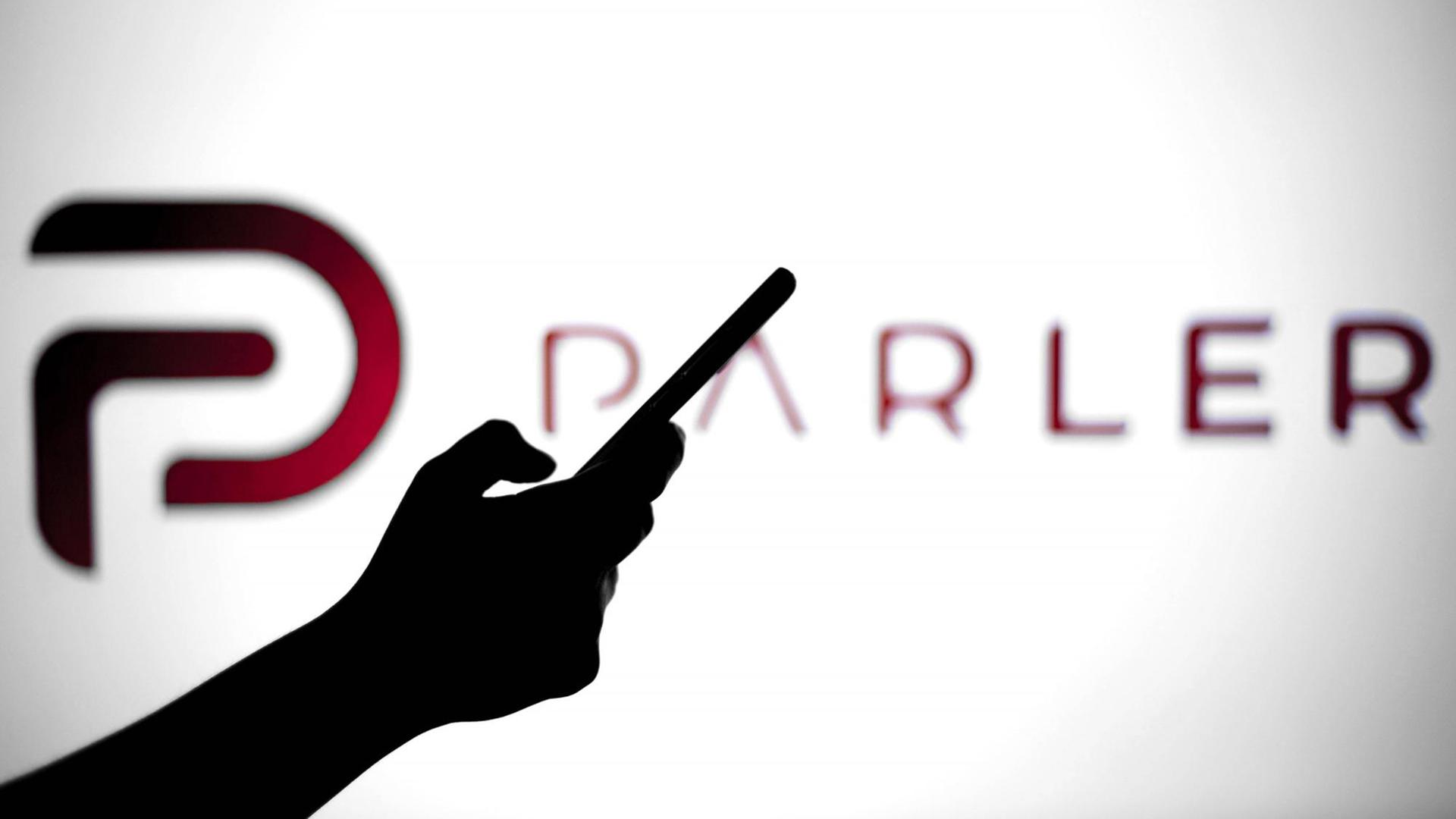 Eine Hand hält ein Smartphone, hinter dem Umriss ist vor leuchtend weißem Hintergrund das Logo von "Parler" zu sehen.