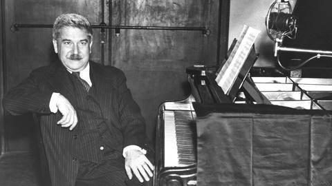 Der Pianist sitzt am Flügel, er hat die Beine überschlagen, der rechte Arm liegt auf der Stuhllehne, er schaut in die Kamera. Er trägt einen Schnauzbart, einen schwarz-weiß gestreiften Anzug mit Weste, ein weißes Hemd und einen schwarz-weiß gestreiften Schlips. er hat kurze, schwarz-graue Haare. Auf dem Notenpult liegen Noten aufgeschlagen, der Deckel des Flügels ist aufgestellt, darunter ist ein Mikrofon positioniert. Der Name des Flügels ist mit schwarzem Tuch abgehängt.