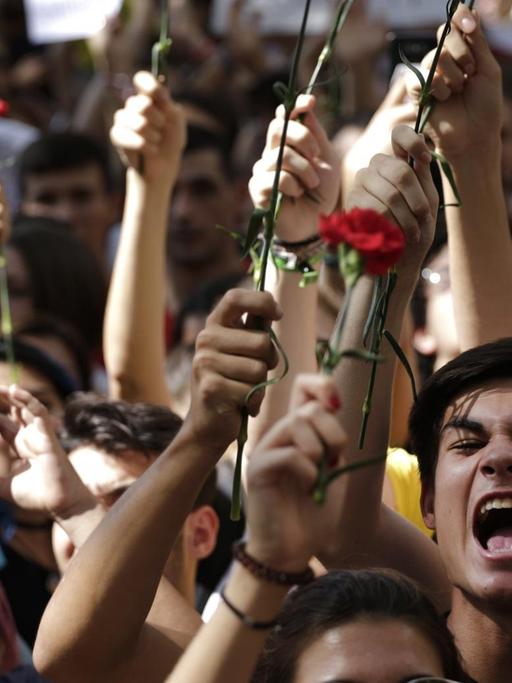 Das Bild zeigt die Köpfe mehrerer Demonstranten, von denen einige etwas rufen. Sie halten die roten Blumen in die Höhe.