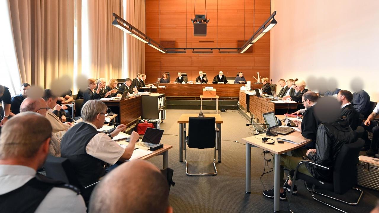 Zu Beginn des Prozesses sitzen elf Angeklagte und Justizbeamte im Landgericht seitlich hinter ihren Anwälten. In der Mitte stehen die Richter und Schöffen.
Zu Beginn des Prozesses sitzen elf Angeklagte und Justizbeamte im Landgericht seitlich hinter ihren Anwälten. In der Mitte stehen die Richter und Schöffen.