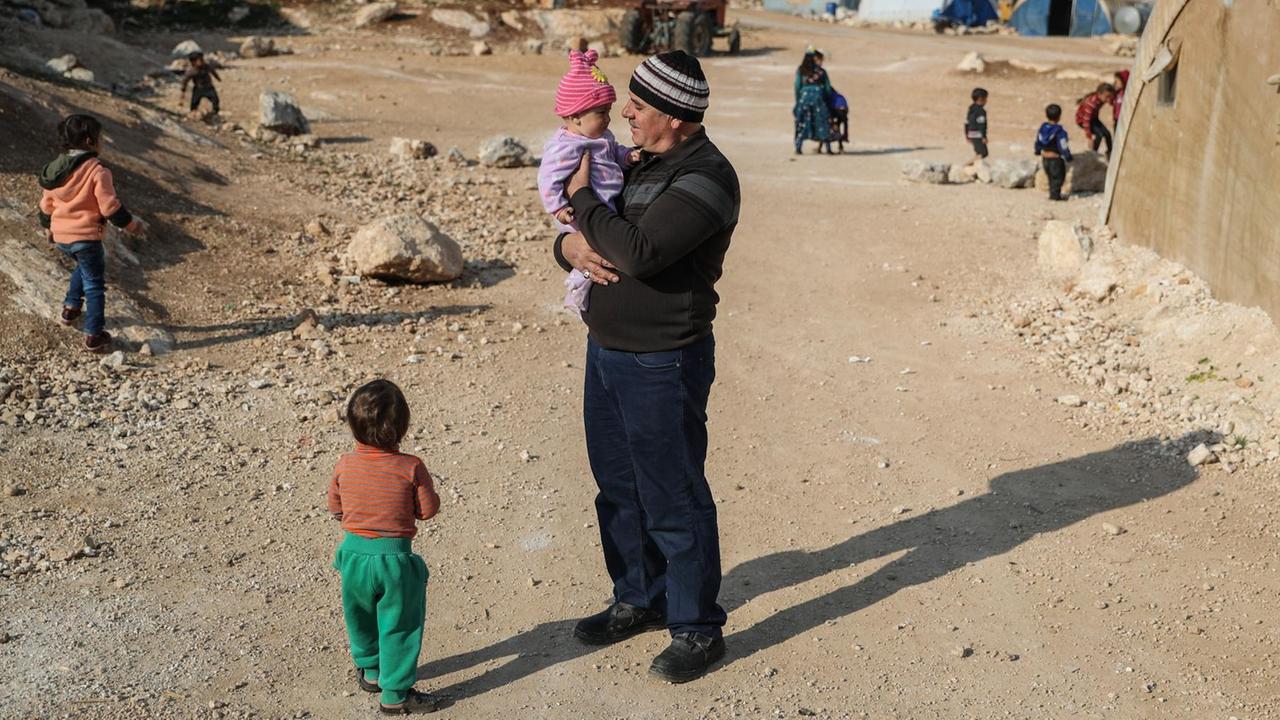 Ein Mann mit Kind auf dem Arm steht in einem staubigen Flüchtlings-Lager in Syrien.