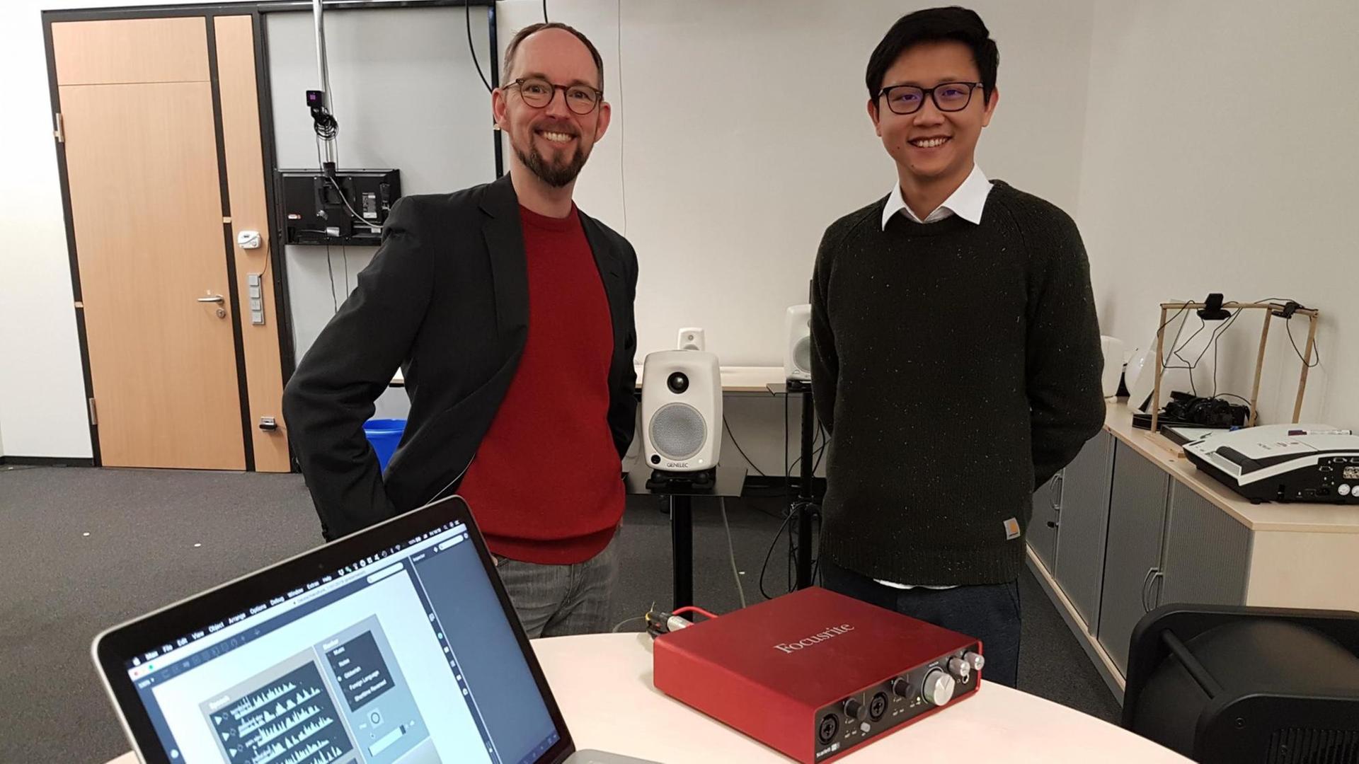 Die Bielefelder Forscher Thomas Hermann und Jiajun Yang stehen nebeneinander in der Mitte eines Arbeitsraumes, im Vordergrund ein Tisch mit einem aufgeklappten Laptop