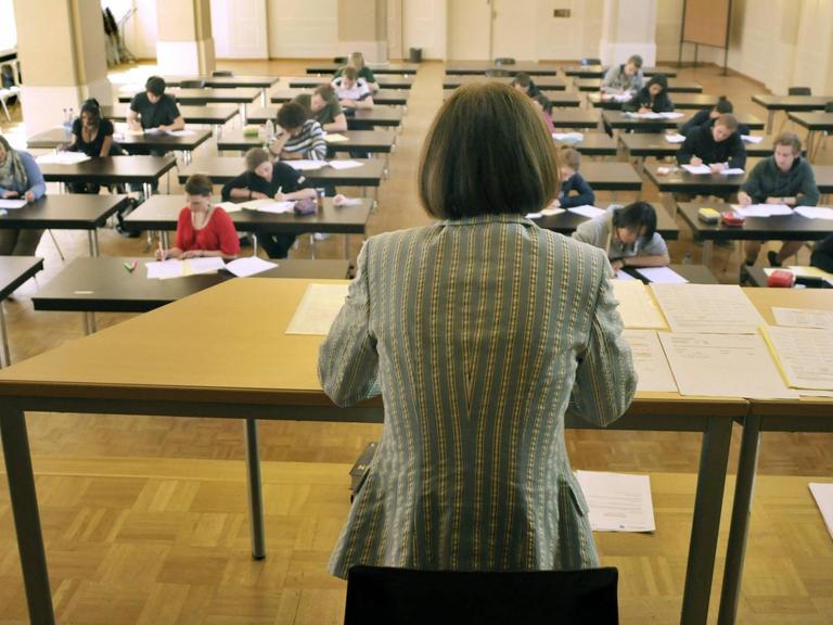 Eine Frau mit dem Rücken zum Betrachter beobachtet Schüler, die an Einzeltischen sitzen und ihr Abitur schreiben.