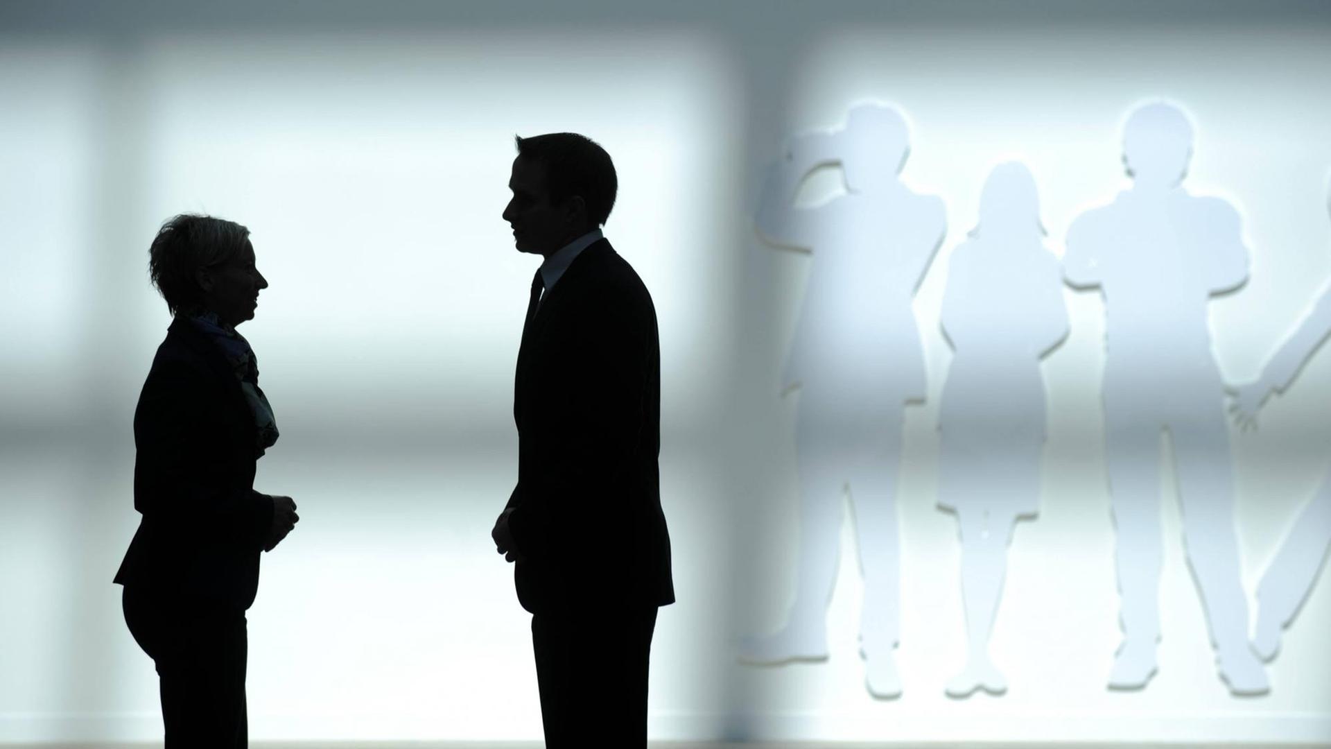 Die Silhouette eines Geschäftsmannes und einer Geschäftsfrau vor einer weißen Wand.