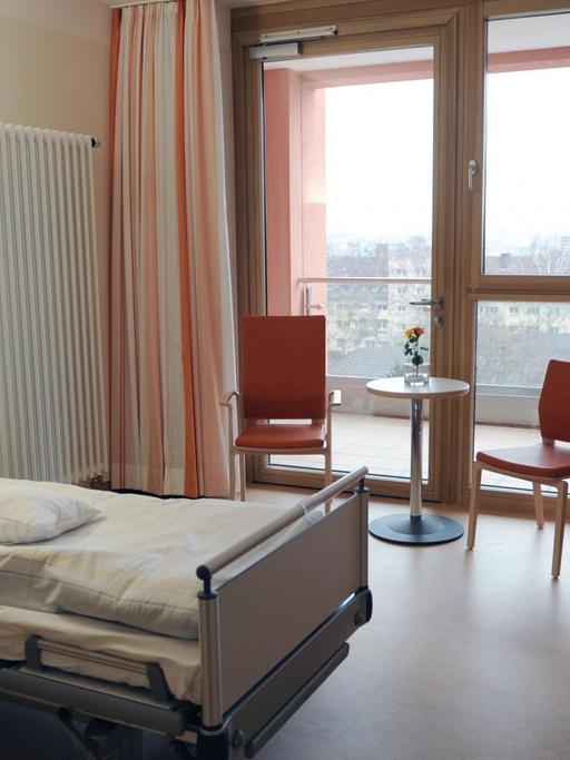Blick in ein Patientenzimmer mit Bett, Tisch, zwei Stühlen und dem Ausblick auf einen Balkon.