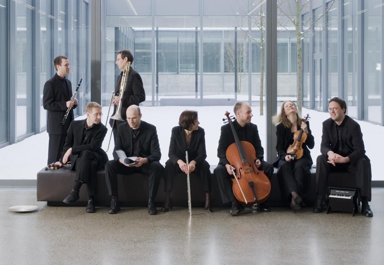 Die Musikerinnen und Musiker des E-Mex Ensemble stehen und sitzen in einer Halle