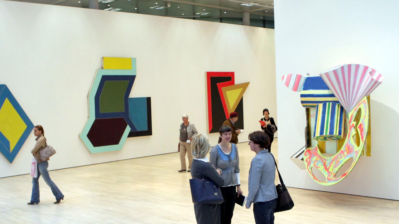 Drei Frauen sprechen im Vordergrund miteinander, an den Wänden im Hintergrund hängen farbenfrohe Kunstwerke