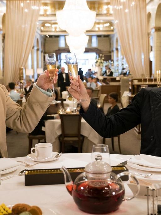 Michael Sheen (li.) als Erzengel Raphael und David Tennant als Dämon Crowley in einer Szene der Comedy-Serie "Good Omens". Die beiden sitzen an einem gedeckten Dinner-Tisch und prosten sich zu.