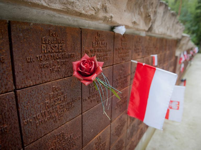 Gedenkstätte für die Opfer des Massenmords von Katyn. Eine polnische Flagge und eine rote Rose stecken an einer Mauer mit eingravierten Namen.